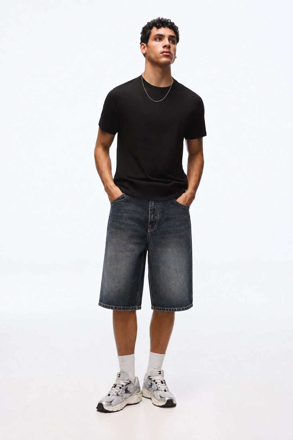 A man modeling a black short-sleeve crewneck t-shirt, dark gray denim shorts, white socks, and white and black sneakers, standing against a plain white studio background.